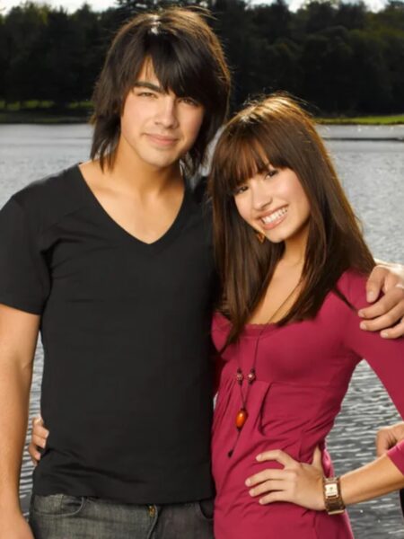 Camp Rock 3 kiedy: Kadr z filmu – młoda dziewczyna w różowej bluzce uśmiecha się i obejmuje chłopaka w czarnej koszulce, stoją razem nad brzegiem jeziora.