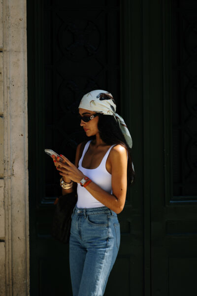 Najmodniejsze kolory jeansów na jesień: Paryż, street style – kobieta w białym body na ramiączkach, do którego dobrała klasyczne jeansy o prostym kroju i apaszkę z nadrukiem, zawiązaną na głowie w stylu retro. Uzupełnieniem są czarne okulary przeciwsłoneczne, złote bransoletki i skórzana torba na ramię.