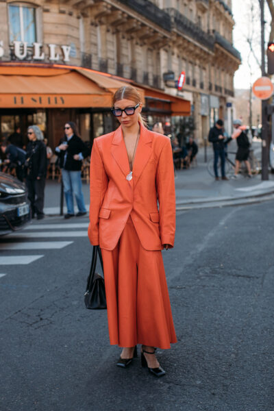 Kobieta uchwycona na ulicach Paryża w stylizacji total look w neutralnych barwach: długa marynarka o pudełkowym kroju w odcieniu beżu, połączona z szerokimi spodniami w tym samym kolorze. Całość dopełniają eleganckie sandały na obcasie, duża torba typu shopper i modne, geometryczne okulary przeciwsłoneczne, które dodają szyku w stylu francuskiej nonszalancji.