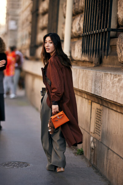 Street style z Paryża: kobieta ubrana w wyrazistą sukienkę midi w odcieniu pomarańczowo-czerwonym, ozdobioną falbanami i geometrycznymi printami. Dopełnieniem stylizacji są metaliczne dodatki – torebka na ramię w srebrnym kolorze i połyskujące sandały na cienkich paskach. Stylizacja łączy w sobie artystyczną ekspresję i modowy eksperyment charakterystyczny dla paryskiego tygodnia mody.