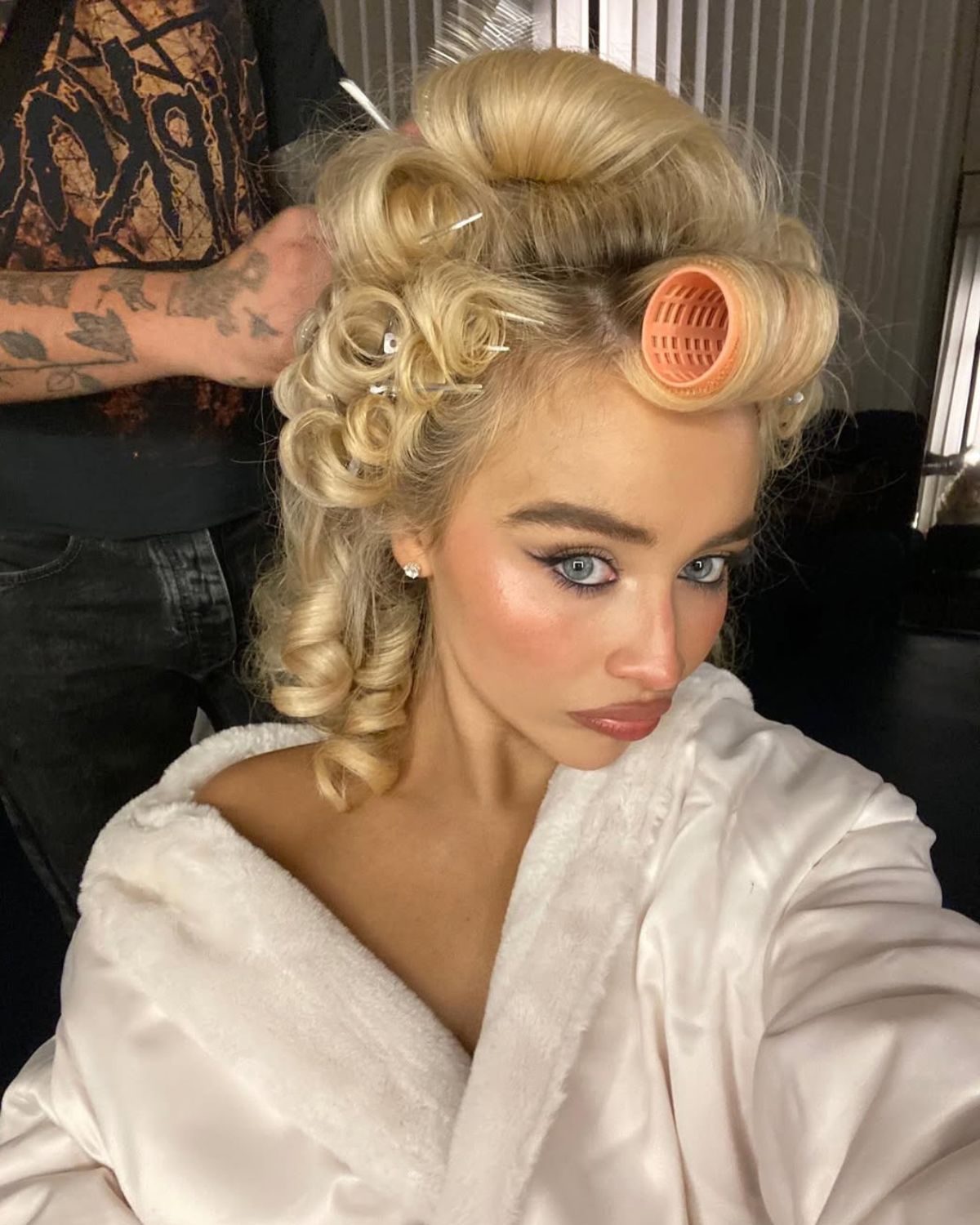 Zdjęcie Sabriny Carpenter, która stosuje drogie, zmysłowe perfumy gwiazd. Jest w trakcie stylizacji, ma wałki na głowie, które zdejmuje jej fryzjer. Ma pełny makijaż. Jest ubrana w kremowy szlafrok. Robi sobie selfie.