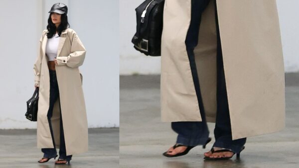 Pedicure Kylie Jenner w długim, oversize’owym, beżowym trenczu narzuconym na biały crop top i ciemne jeansy z rozszerzaną nogawką. Stylizację uzupełnia czarna skórzana torba, czarne japonki na obcasie oraz czapka z daszkiem z czarnej skóry. Ujęcie wykonane w parkingu podziemnym.