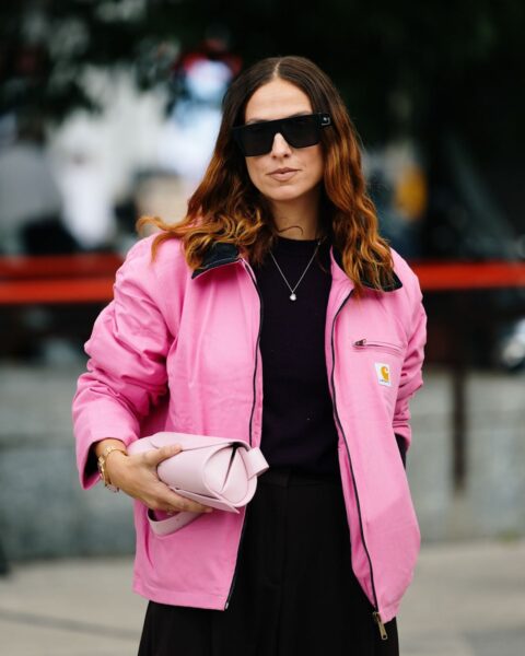 Modna kobieta w wyrazistej, różowej kurtce typu worker jacket z logo Carhartt. Kurtka w kolorze fuksji zestawiona jest z czarnym topem i spódnicą lub spodniami, tworząc kontrastowy i nowoczesny look. Uzupełnieniem stylizacji są duże, prostokątne okulary przeciwsłoneczne, delikatny naszyjnik z zawieszką oraz jasnoróżowa, designerska torebka-saszetka trzymana w dłoni. Inspiracja odważnym kolorem i połączeniem streetwearu z klasą.