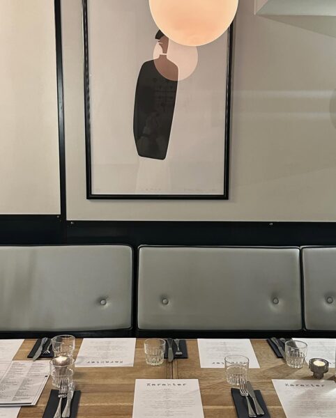 Wnętrze restauracji z elegancko nakrytym stołem – menu, sztućce i szklanki ułożone dla kilku osób, w tle jasna tapicerowana ławka oraz nowoczesny obraz na ścianie.