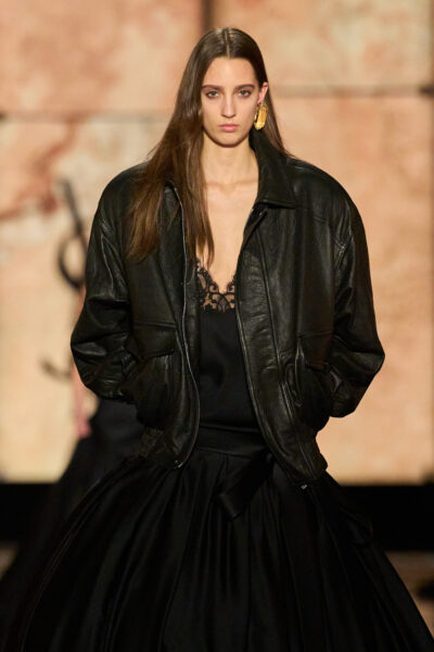 Modelka na pokazie Saint Laurent FW25 w klasycznej czarnej, skórzanej bomberce oversize zestawionej z elegancką, czarną suknią o objętościowym kroju i koronkowym topie. Stylizacja łączy elementy codzienne z haute couture.
