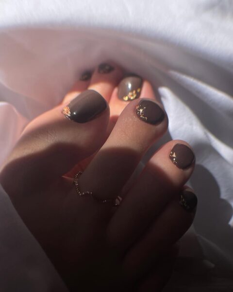 Brązowy pedicure z subtelnymi złotymi płatkami wkomponowanymi przy krawędziach paznokci – jesienny, nastrojowy manicure stóp w klimacie „quiet luxury”, podkreślający naturalne ciepło barw.