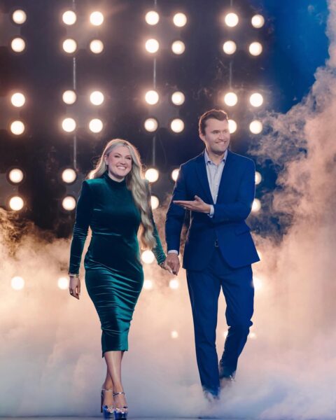 Para w eleganckich strojach wychodzi na scenę wśród świateł i dymu, trzymając się za ręce.