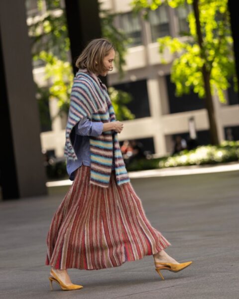 Kobieta w stylizacji street style sfotografowana przed pokazem Toteme podczas New York Fashion Week SS26. Ma na sobie długą, plisowaną spódnicę w ciepłe, pionowe pasy (odcienie czerwieni, brązu i beżu), błękitną koszulę oraz narzucony na ramiona gruby, dzianinowy szal w kolorowe pasy. Stylizację uzupełniają żółte czółenka na obcasie.