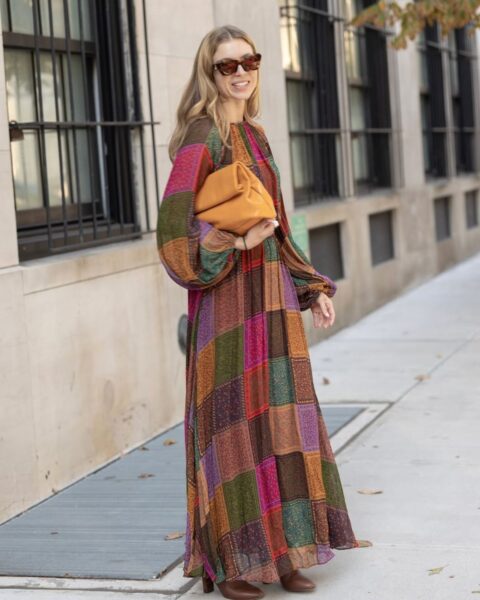 Kobieta w stylizacji street style sfotografowana przed pokazem Ulla Johnson podczas New York Fashion Week SS26. Ma na sobie długą, zwiewną sukienkę maxi z bufiastymi rękawami, uszytą z kolorowych, patchworkowych paneli w różnych, etnicznych/paisley wzorach (odcienie brązu, zieleni, czerwieni, fuksji). W ręku trzyma dużą, koniakową torebkę-kopertówkę (typu pouch) i nosi brązowe botki oraz okulary przeciwsłoneczne.