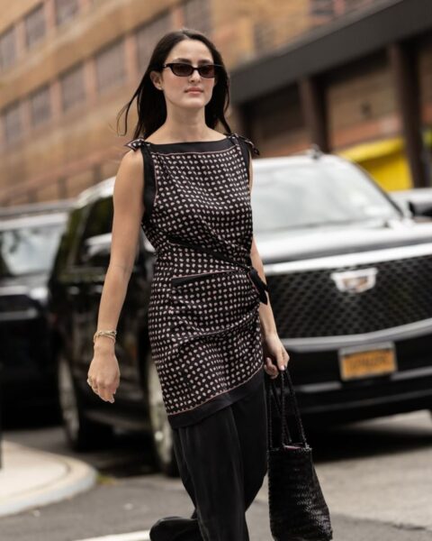 Kobieta w stylizacji street style sfotografowana przed pokazem Area podczas New York Fashion Week SS26. Ma na sobie asymetryczną tunikę z wiązaniami na ramionach i drobnym, geometrycznym wzorem (czarno-różowym) oraz szerokie, czarne spodnie. W ręku trzyma czarną, plecioną torebkę typu koszyk, na stopach ma czerwone klapki, a oczy zakrywają prostokątne okulary przeciwsłoneczne.