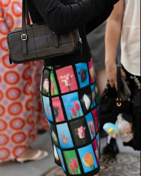 Detal stylizacji street style sfotografowany podczas New York Fashion Week SS26. Kobieta ma na sobie czarną, dopasowaną bluzkę oraz spódnicę mini z wyrazistym, wielobarwnym wzorem w tulipany ujęte w kwadratowe, czarne ramy. W ręku trzyma czarną, prostokątną torebkę na ramię z klamrą.