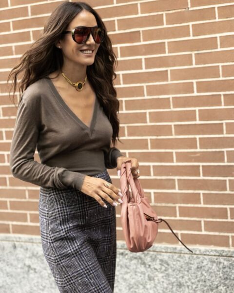 Kobieta w eleganckiej stylizacji street style sfotografowana podczas Milan Fashion Week SS2026 na tle ceglanej ściany. Ma na sobie brązowy sweter z dekoltem w serek, spodnie w szaro-niebieską kratę (pepitkę/księcia Walii), wyrazisty złoty naszyjnik typu choker z okrągłym wisiorem oraz duże, brązowe okulary przeciwsłoneczne. W ręce trzyma różową, marszczoną torebkę.