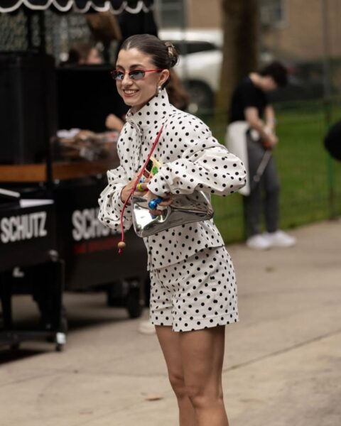 Kobieta w stylizacji street style sfotografowana przed pokazem Area podczas New York Fashion Week SS26, uśmiecha się do koleżanki. Ma na sobie dwuczęściowy komplet (koszulę ze stójką i szorty) w biało-czarne, drobne grochy. Stylizację uzupełnia metaliczna, srebrna torebka typu bagietka noszona w pasie, czarne klapki na obcasie oraz okulary przeciwsłoneczne w czerwonej oprawce.