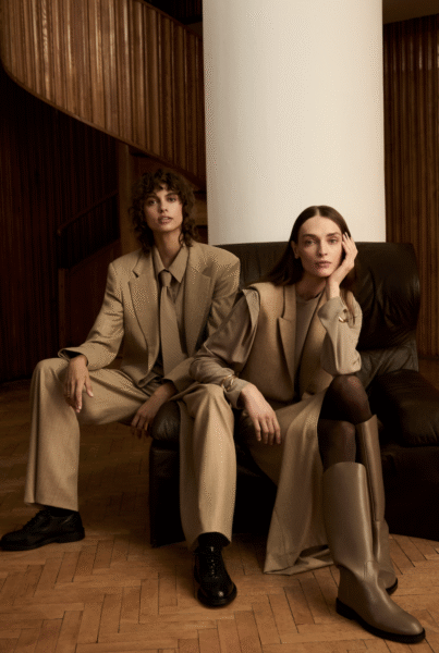 Dwie modelki w stylizacjach z kolekcji JEMIOŁ AW25 siedzące na skórzanym fotelu. Jedna w oversize’owym garniturze w odcieniu cappuccino z szerokimi spodniami i krawatem, druga w długim bezrękawnym płaszczu w karmelowym kolorze, zestawionym z kozakami do kolan. Elegancja w klimacie power dressing.