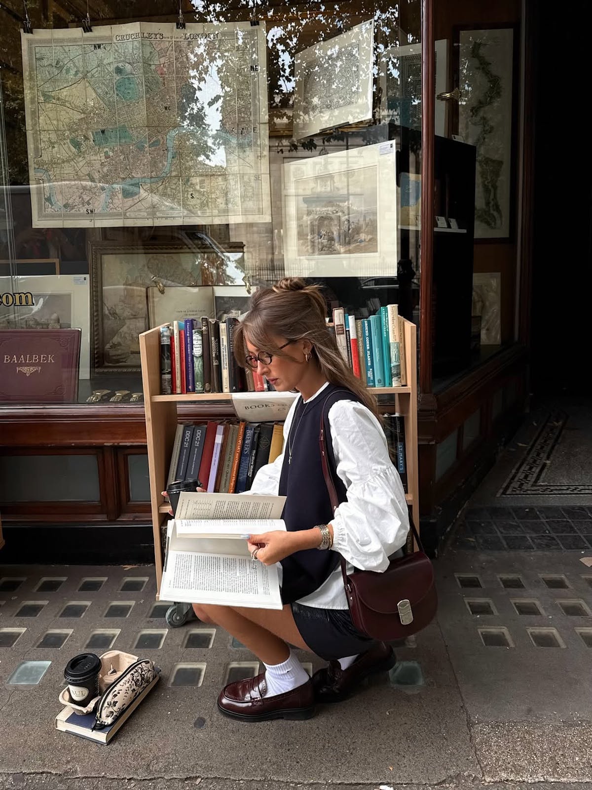 Osoba siedząca przed antykwariatem, przeglądająca książki na zewnętrznym regale – jesienna scena czytelnicza z książką i kawą