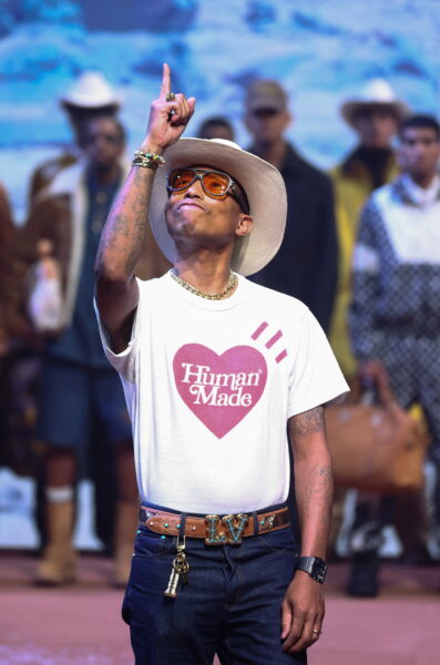 Najciekawsze współprace mody i muzyki: Pharrell Williams w białym T-shircie Human Made i kowbojskim kapeluszu podczas swojego debiutanckiego pokazu dla Louis Vuitton, unoszący rękę w geście triumfu, na tle modeli prezentujących kolekcję męską.