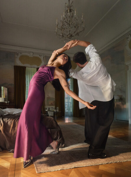 Stylizowane zdjęcie pary w eleganckim wnętrzu – kobieta w satynowej, śliwkowej sukni tańczy z mężczyzną w białej koszuli i czarnych spodniach. Jej ciało odchylone jest w teatralnym geście, a srebrne buty na obcasie połyskują w świetle wpadającym przez okna. Scena przypomina kadr z filmu o miłości i namiętności, rozgrywającym się w pałacowych wnętrzach.