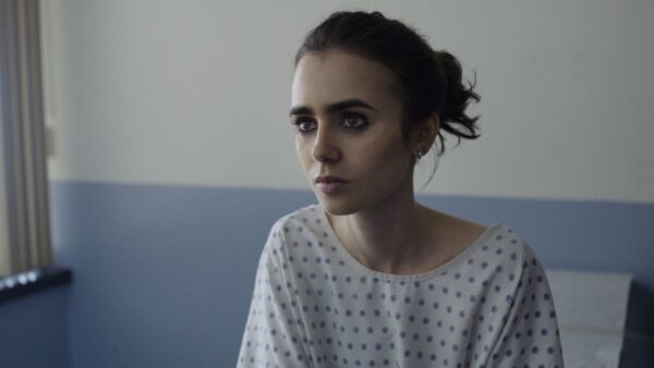 Kadr z filmu „Aż do kości” z Lily Collins – aktorka w roli Ellen, młodej kobiety zmagającej się z anoreksją. Ubrana w szpitalną koszulę, siedzi na łóżku i spogląda poważnie w bok. Zdjęcie kontrastuje z jej glamour’owymi wyjściami na czerwone dywany.