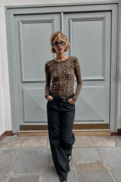 Modelka w dopasowanym body w panterkę i ciemnych, szerokich jeansach o kroju relaxed fit, pozująca na tle klasycznych, szarych drzwi. Stylizacja utrzymana w duchu minimalistycznego street style’u z lat 2000., podkreślona czarnym paskiem i okularami o kocim kształcie.
