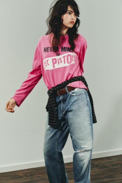 Młoda kobieta w różowej bluzie z nadrukiem „Never Mind the Sex Pistols” i przetartych jeansach typu boyfriend, zawiązaną w pasie pasiastą bluzą. Stylizacja inspirowana punkową energią i modą vintage.
