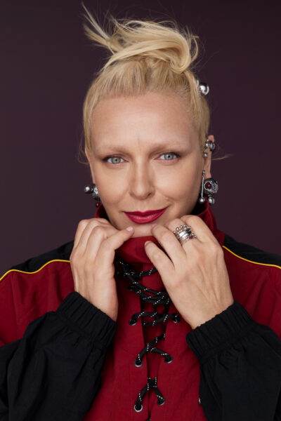 Portret kobiety o blond włosach upiętych w nowoczesny kok, ubranej w bordowo-czarną kurtkę z wiązaniem. Stylizacja uzupełniona dużymi, rzeźbiarskimi kolczykami i pierścionkami o srebrnym połysku. Makijaż z mocnym akcentem ust w kolorze fuksji i spokojnym spojrzeniem podkreśla siłę kobiecej ekspresji.