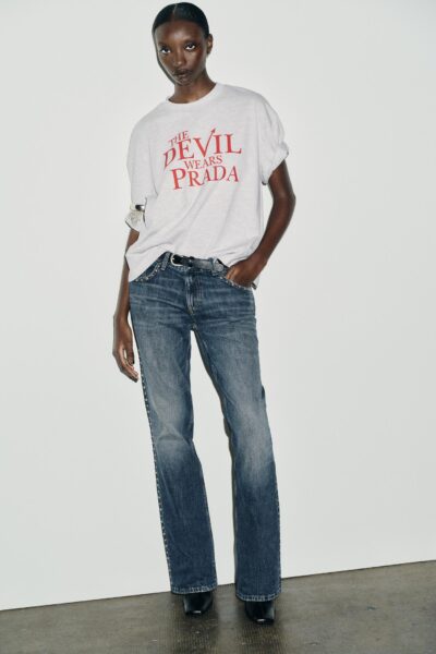 Kobieta w jasnoszarej koszulce z czerwonym napisem „The Devil Wears Prada” i prostych jeansach, pozująca na minimalistycznym, białym tle. Look inspirowany kulturą pop i ironią mody lat 2000.