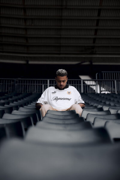 Portret mężczyzny siedzącego samotnie w rzędzie pustych krzeseł stadionowych, w białej koszulce z logo PUMA i napisem „Represent”, w nastrojowej, przygaszonej tonacji zdjęcia.