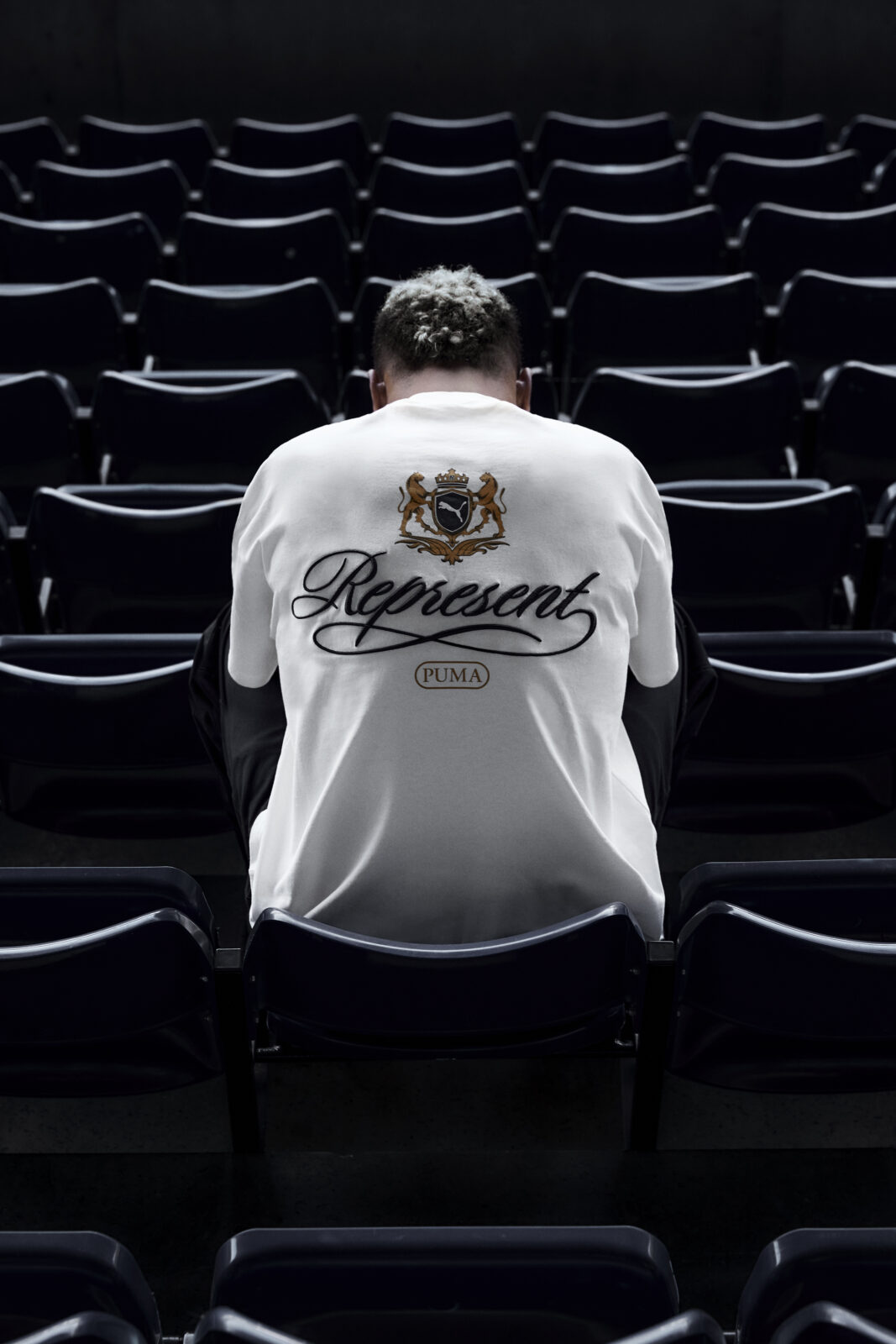 PUMA x Represent: Zdjęcie od tyłu – mężczyzna siedzi na stadionowych krzesłach, w białej koszulce z herbem i logotypem PUMA x Represent wyszytym z tyłu. Ujęcie podkreśla streetwearowy charakter kampanii.