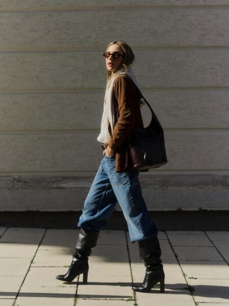 10 elementów garderoby, które budują status fashion girl. Kobieta w stylizacji opartej na kontrastach: luźny dół (dżinsy) i dopasowane, wysokie kozaki na obcasie. Marynarka w kolorze karmelu. Elementy garderoby, które budują status fashion girl: okulary w modnej oprawce i duża, pojemna torba typu worek, dodające całości nonszalancji.