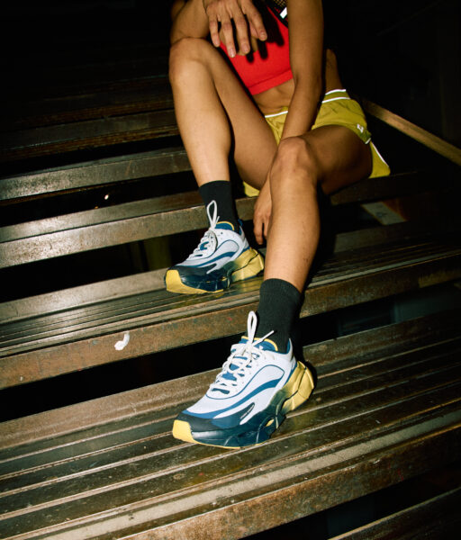 Kobieta siedząca na stalowych schodach, ubrana w czerwony sportowy top, żółte szorty i sneakersy Under Armour HALO. Gestem poprawia okulary, a tło z ciemnymi konstrukcjami przemysłowymi nadaje scenie surowy, streetwearowy klimat.