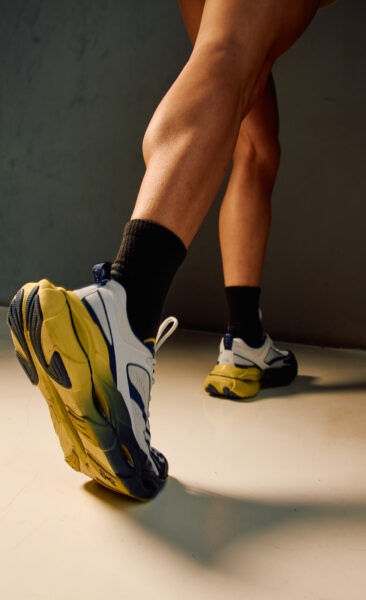 Zbliżenie na buty sportowe w kolorach bieli, granatu i żółci z masywną, futurystyczną podeszwą. Zdjęcie oddaje dynamikę ruchu, skupiając się na kroku w biegu – detal nawiązujący do miejskiej energii i mocy sportu.