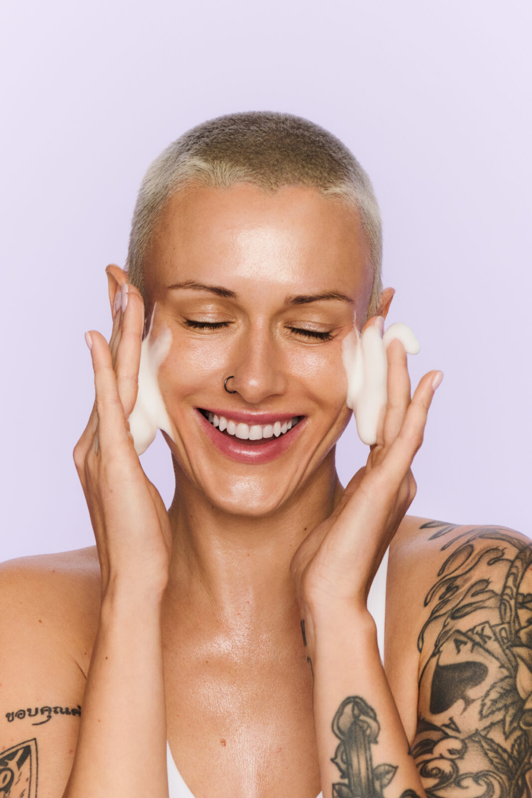 KLOO marka: Modelka z krótkimi włosami i kolczykiem w nosie uśmiecha się szeroko, rozprowadzając na twarzy delikatną piankę oczyszczającą KLOO. Ujęcie pełne energii i świeżości, z lawendowym tłem w estetyce wellness.