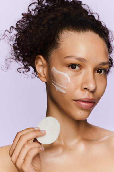 Kobieta o naturalnej urodzie z upiętymi, kręconymi włosami nakłada krem pielęgnacyjny na policzek, trzymając w dłoni płatek kosmetyczny. Skóra rozświetlona, zdrowa i gładka, na tle delikatnego, lawendowego tła – kadr oddający ideę świadomej pielęgnacji KLOO.