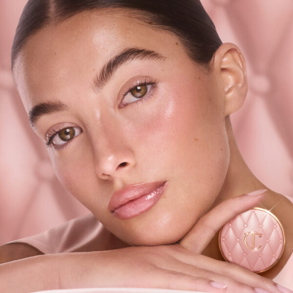 Portret kobiety o naturalnym makijażu i błyszczącej cerze, która delikatnie opiera głowę na dłoni, trzymając różowy kompakt Charlotte Tilbury. Tło w kolorze nude podkreśla subtelny blask i luksusowy charakter produktu do makijażu.
