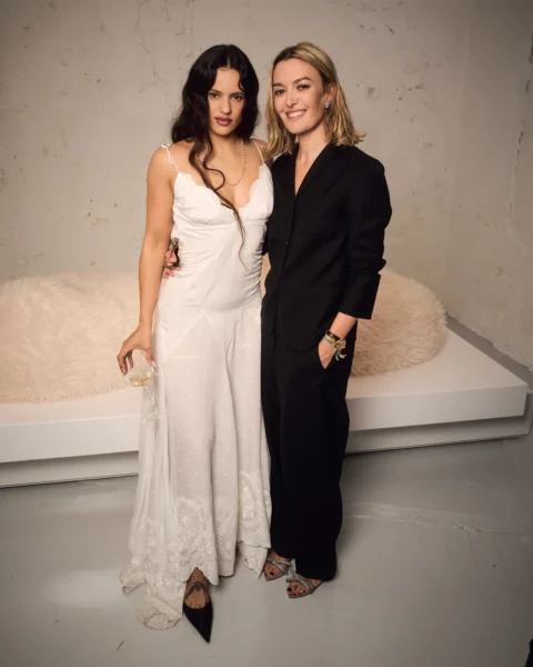 Kolekcja Zara 50 Creators: Rosalía i Marta Ortega Pérez na evencie ZARA – Rosalía w długiej białej sukni na cienkich ramiączkach z koronkowym wykończeniem i czarnych szpilkach oraz Marta Ortega Pérez w czarnym garniturze i sandałach na obcasie, pozują wspólnie do zdjęcia.