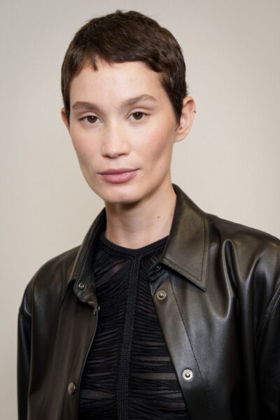 Portret kobiety z krótką fryzurą pixie, o naturalnym makijażu i subtelnym wyrazie twarzy. Ma na sobie czarną, skórzaną kurtkę i top z transparentnymi wstawkami, co nadaje jej stylizacji minimalistycznego, nowoczesnego charakteru.