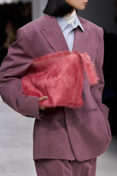 Torebki wiosna–lato 2026: Zbliżenie na modelkę w oversize’owym garniturze w odcieniu przygaszonej purpury, trzymającą w dłoni kopertówkę z różowego sztucznego futra. Miękka faktura torebki kontrastuje z męskim krojem garnituru, tworząc look w duchu playful power dressing.