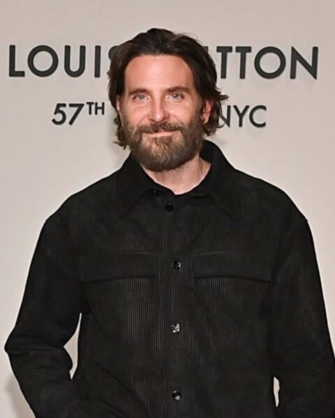 Zdjęcie przedstawia aktora Bradleya Coopera stojącego na tle jasnej ściany z logo "LOUIS VUITTON 57TH NYC". Aktor ma długą, gęstą brodę i włosy zaczesane do tyłu. Ubrany jest w czarną, zapinaną koszulę lub kurtkę z prążkowanej tkaniny z dwiema kieszeniami na piersi. Patrzy prosto w obiektyw z lekkim uśmiechem.