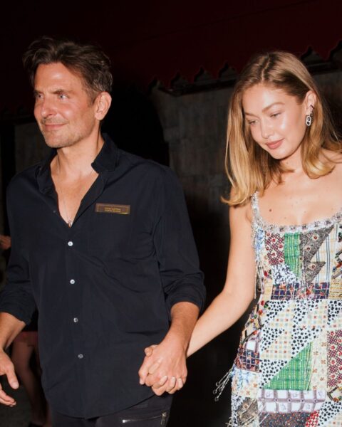 Zdjęcie przedstawia aktora Bradleya Coopera i modelkę Gigi Hadid, idących obok siebie i trzymających się za ręce w nocy, prawdopodobnie wychodzących z jakiegoś lokalu. Bradley Cooper jest ubrany w czarną koszulę i czarne spodnie, patrzy przed siebie z lekkim uśmiechem. Gigi Hadid ma na sobie długą sukienkę na ramiączkach, uszytą z tkaniny przypominającej patchwork w różne wzory i kolory. Patrzy w dół i uśmiecha się. W tle widać ciemny budynek i zamazane postacie.