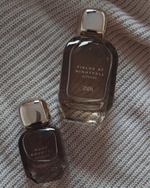 Dwie butelki intensywnych perfum Zara leżące na szarym, dzianinowym tle. Większy flakon to Fields at Nightfall Intense, a mniejszy to Nude Bouquet Intense. Oba mają szklane, dymione butelki w kolorze brązowo-szarym i metaliczne, złote lub srebrne nakrętki.