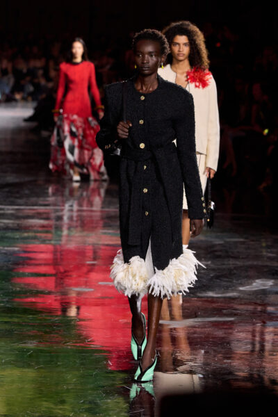 Modelka w czarnym, tweedowym płaszczu o złotych guzikach, wykończonym białymi piórami u dołu. Stylizacja uzupełniona błyszczącymi kolczykami i miętowymi butami to nowoczesne spojrzenie na kod Chanel — elegancję, która nie boi się kontrastów.