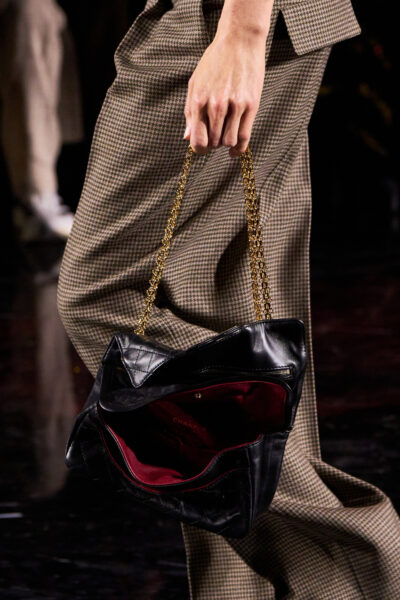 Detal: klasyczna czarna skórzana torba Chanel o miękkiej konstrukcji, z burgundową podszewką i złotym łańcuszkiem. Modelka w garniturze w kratkę prezentuje współczesną interpretację paryskiej elegancji z nutą nonszalancji.