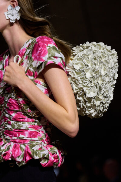 Torebka w formie dużego kwiatowego bukietu z białych, przestrzennych aplikacji noszona na ramieniu przez modelkę w kwiecistej bluzce z różowym printem. Stylizacja celebruje romantyzm haute couture i teatralny charakter marki.