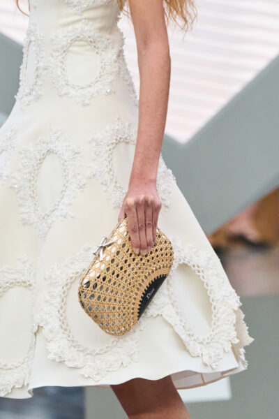 Zbliżenie na kremową, haftowaną suknię z barokowymi aplikacjami i torebkę Dior w formie wiklinowej kopertówki z delikatną kokardką. Zestaw łączy klasyczną francuską elegancję z letnim rzemiosłem couture, nawiązując do estetyki ogrodów i światła.