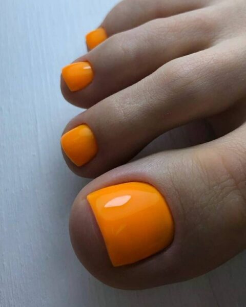 Zbliżenie na stopę z intensywnym, błyszczącym dyniowym pedicure w soczystym, neonowym odcieniu pomarańczu. Pedicure idealnie pasuje do letnich sandałów i jesiennych stylizacji, podkreślając opaleniznę. Kolor jest jednolity, a paznokcie są starannie opiłowane.