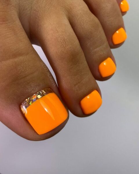 Zbliżenie na stopę z letnim pedicure w energetycznym, neonowym odcieniu pomarańczy. Paznokieć dużego palca u stopy ozdobiony jest dodatkowo delikatnym, odwróconym french manicure, gdzie linia u nasady paznokcia (przy skórkach) została podkreślona błyszczącymi, holograficznymi drobinkami brokatu. Reszta paznokci ma jednolity, lśniący kolor.
