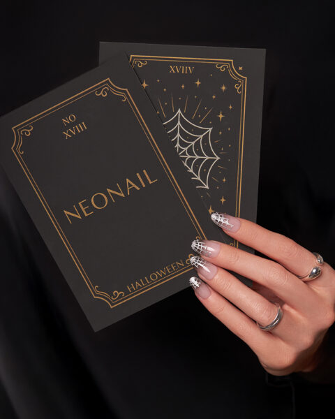 Zdjęcie dłoni z manicure NEONAIL – paznokcie ozdobione srebrnymi pajęczynami na tle delikatnego, mlecznego ombré. W dłoni karty inspirowane tarotem z napisem „Halloween Collection” i logotypem NEONAIL w złotych detalach.