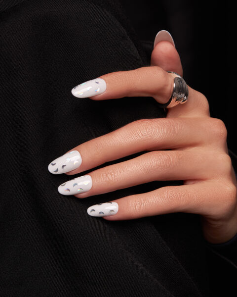 Zbliżenie dłoni z białym manicure i holograficznymi naklejkami w kształcie półksiężyców. Lśniące elementy odbijają światło, tworząc efekt ruchomej poświaty – elegancki i nowoczesny wzór w stylu „celestial nails”.