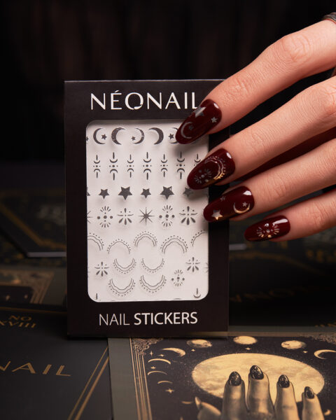 Dłoń z bordowym, błyszczącym manicure NEONAIL dekorowanym srebrnymi motywami gwiazd i księżyców trzyma opakowanie naklejek do paznokci „Nail Stickers” z kolekcji o kosmicznym charakterze. Tło stanowią ilustracje w złocie i czerni, podkreślające magiczny klimat stylizacji.
