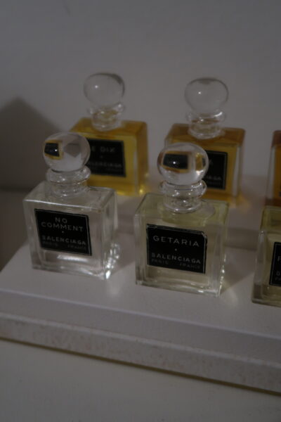 Perfumy Balenciaga: Zbliżenie na flakony Balenciaga Getaria i No Comment – dwa przezroczyste, szklane flakoniki perfum z klasycznymi okrągłymi zatyczkami, w których widoczne są lekkie płyny zapachowe, ustawione na białej podstawce w towarzystwie pozostałych miniaturek.